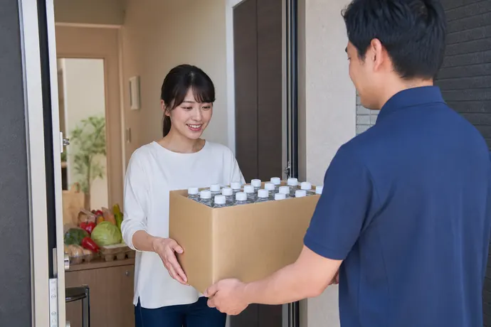 自宅の玄関で宅配スタッフからペットボトルの水を受け取る女性