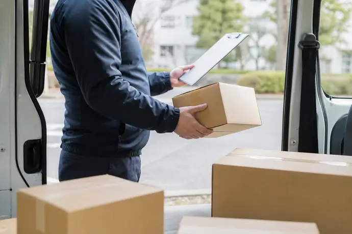 軽貨物ドライバーが安全な停車場所で一度立ち止まり、荷物と置き配指定を確認している場面。急いで走る様子や危険運転、文字やロゴは入れない。