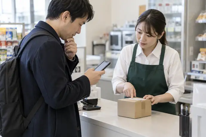 コンビニのレジ前でスマートフォンの受取通知を確認しながら荷物を受け取る様子