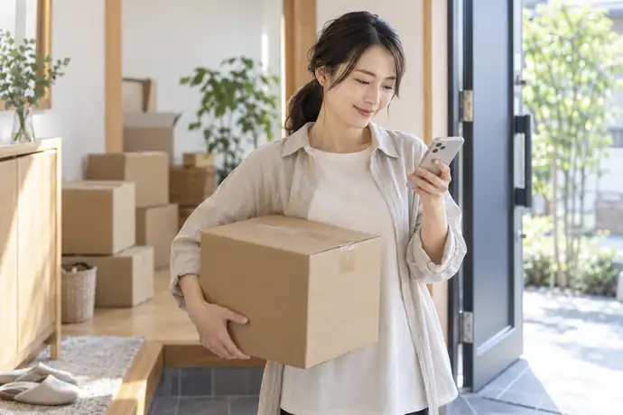引っ越し後の玄関で小包とスマートフォンを手に転送先を確認する受取人